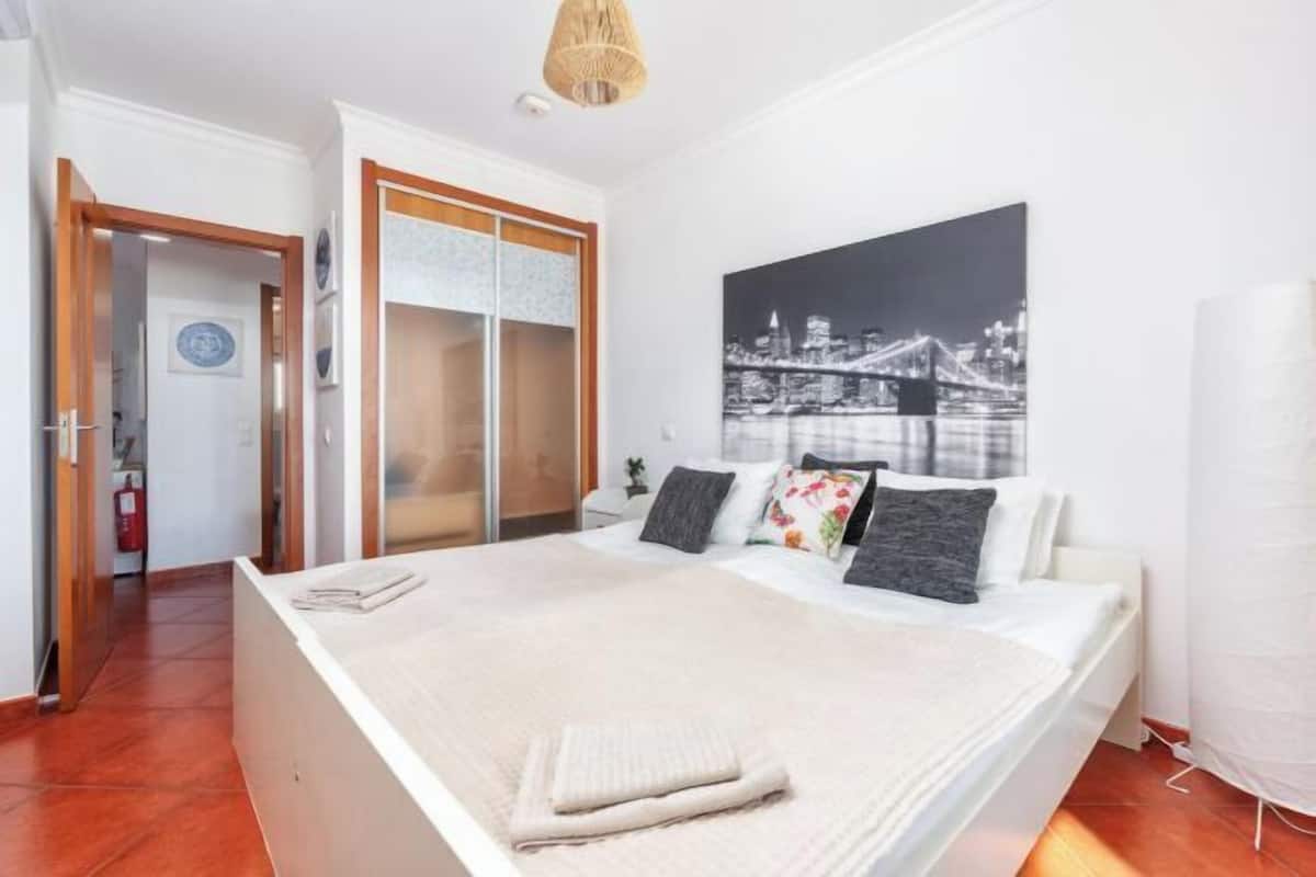 A bedroom with a bed, a cityscape painting, a wardrobe, and a door leading to another room.