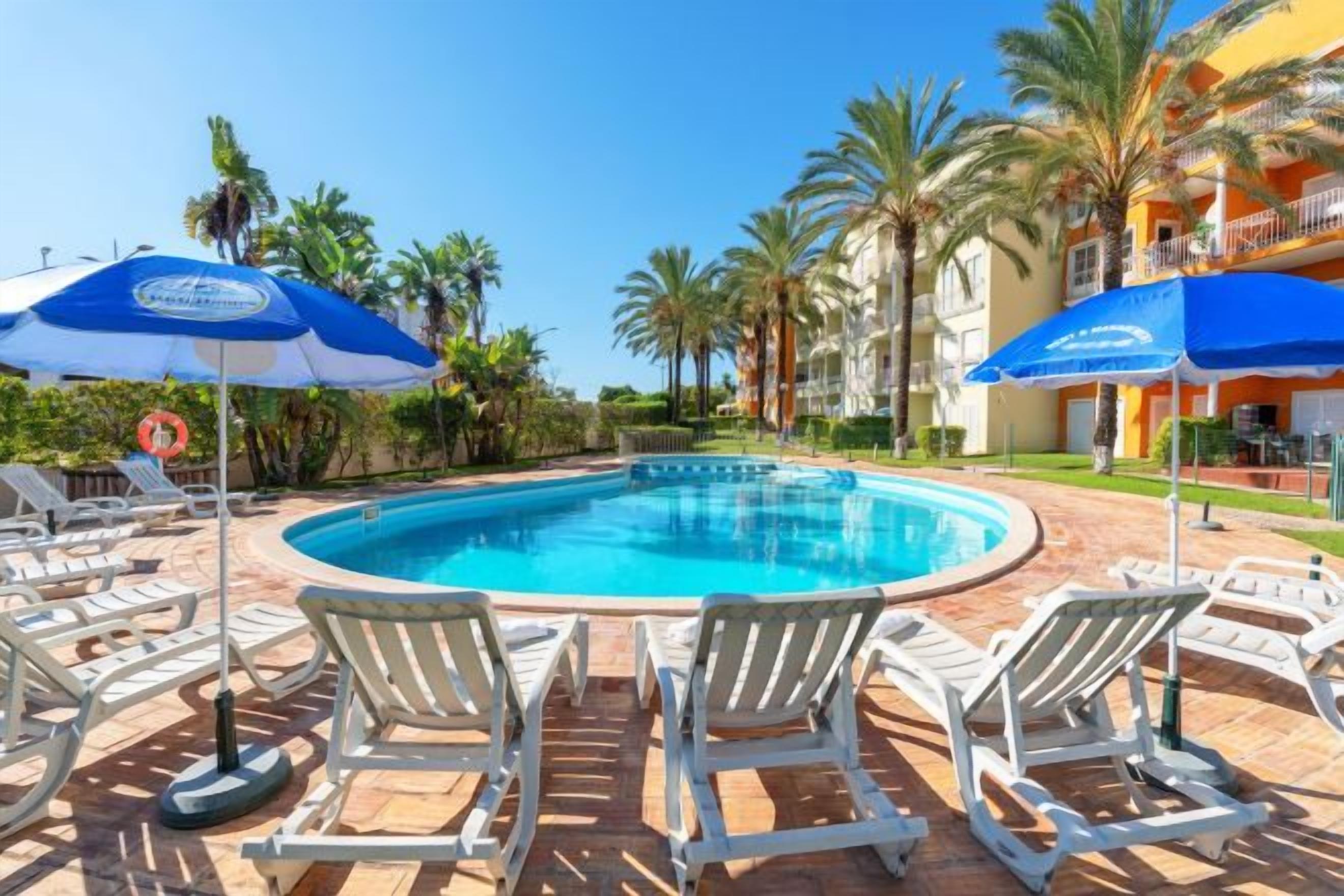 A swimming pool with lounge chairs and umbrellas, surrounded by palm trees and a building with balconies.