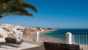 A seaside balcony with two wooden benches, overlooking a beach with a clear blue sky and a coastal town in the background. - BnB Homes Apartment with cozy Terrace & Swimming pool near the heart city (Albufeira)