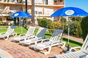 Ein Poolbereich mit Liegestühlen und Sonnenschirmen, ein Gebäude mit Balkonen und eine gepflegte Rasenfläche.