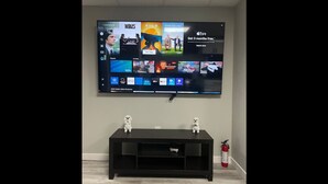 A flat-screen TV mounted on a wall above a black entertainment unit with two small white figurines on top. - Cutee Home in Prairie Pointe (Winnipeg)