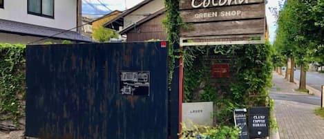 A rustic wooden sign with 'Cotohama Green Shop' written on it, a closed blue metal gate, and a small signboard with 'CLAMP COFFEE' written on it.