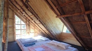 Een kleine, rustieke slaapkamer op zolder, met een schuine kap, houten balken en een enkel bed met matras en kussens.