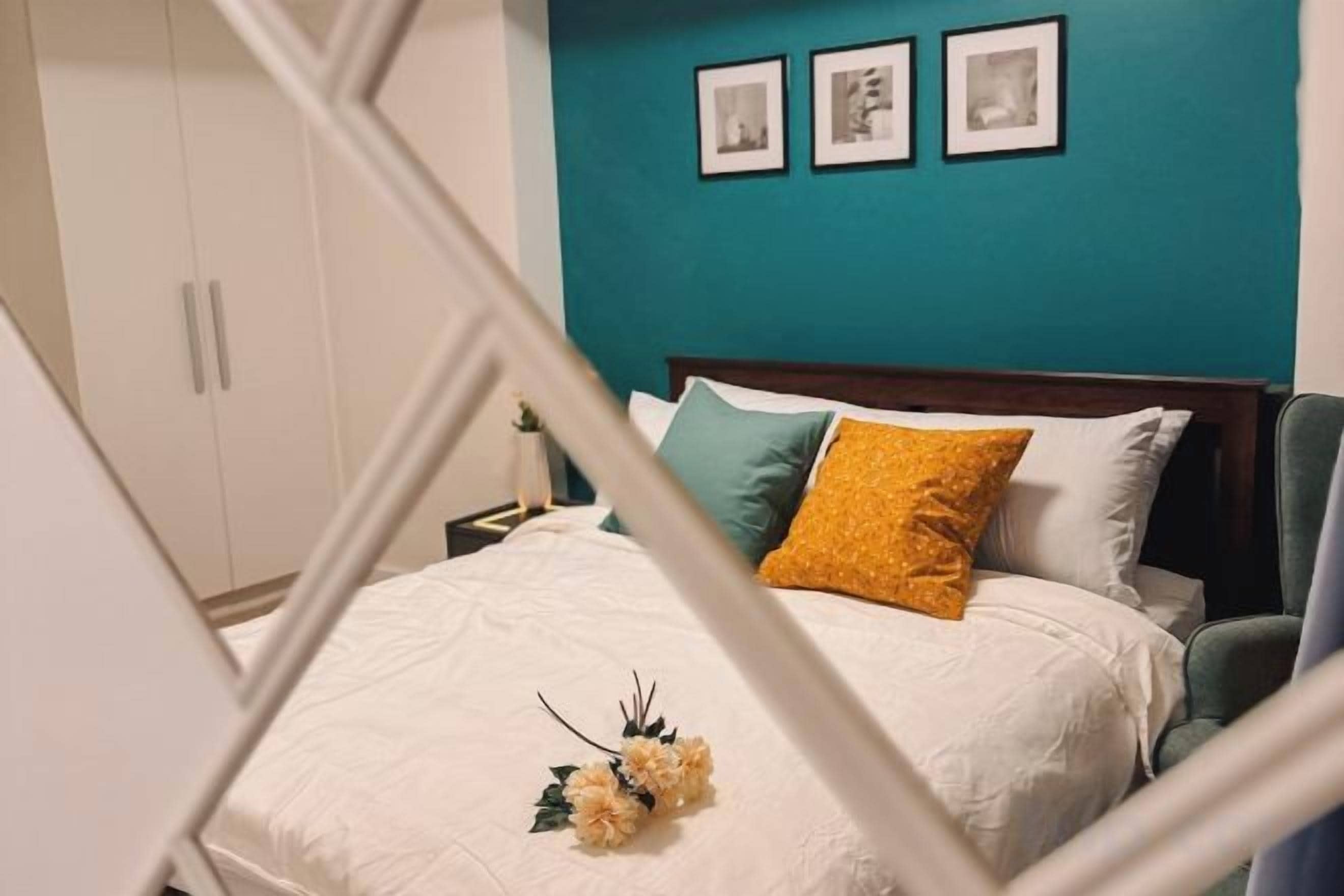 Una cama con edredón blanco, almohada amarilla y decoración floral. Encima de la cama, hay tres cuadros enmarcados en la pared.
