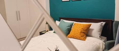 Una cama con edredón blanco, almohada amarilla y decoración floral. Encima de la cama, hay tres cuadros enmarcados en la pared.