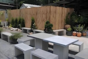 Mobiliario exterior con mesas y bancos de hormigón, rodeado de plantas en macetas y una valla de bambú.