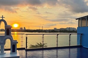 A sunset view from a balcony with a railing, a small building with a cross, and a cityscape in the distance. - Santorini 3000sqft Townvilla, rooftop BBQ, 5BR, 13pax (Puchong)