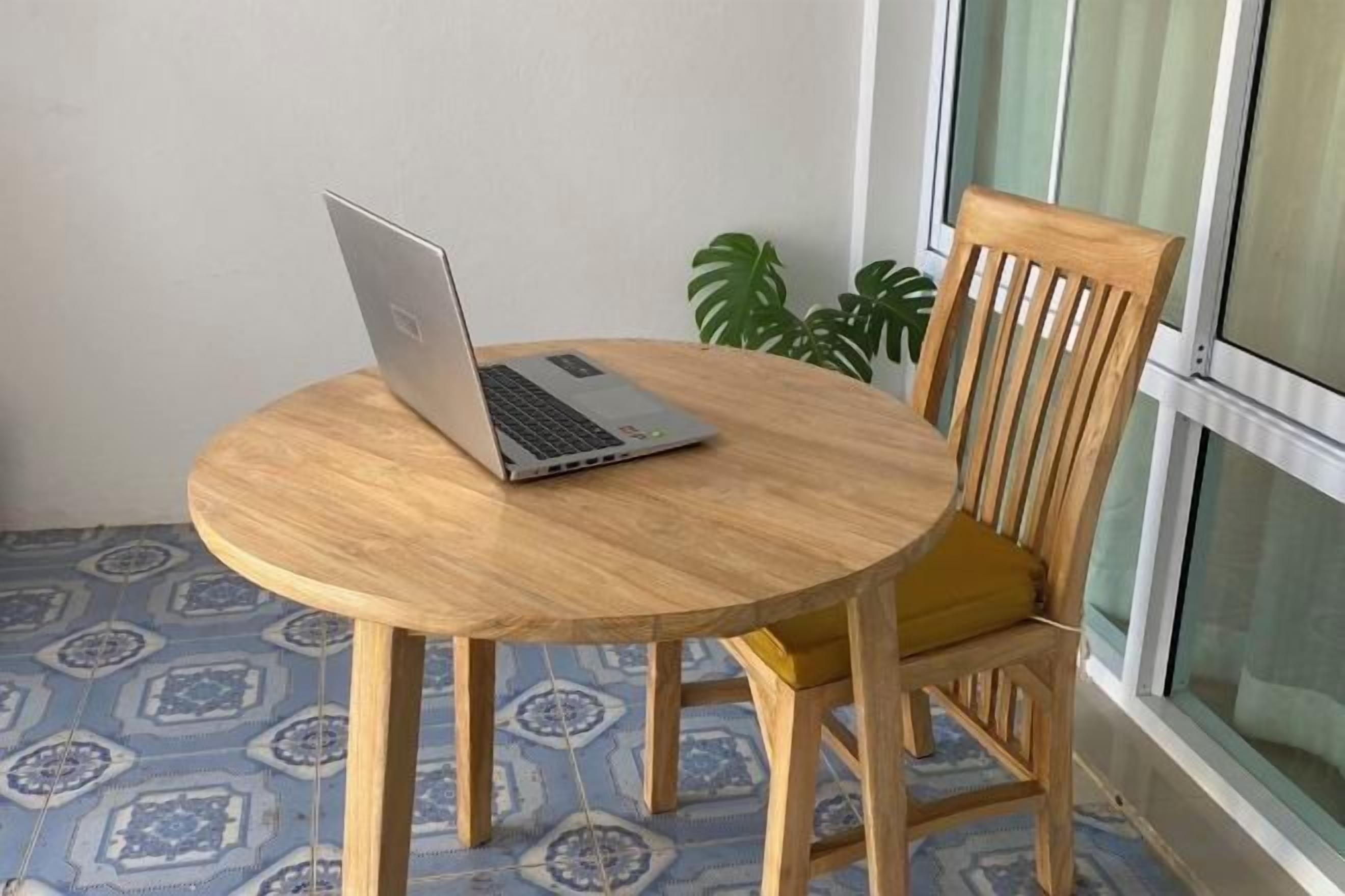 A round wooden table with a laptop on it, a wooden chair with a yellow cushion, and a patterned floor.