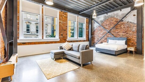 A modern bedroom with a large bed, a gray sofa, and a view of a brick wall. - Modern Studio + Prime Location by Zodiak Stays (Auckland)