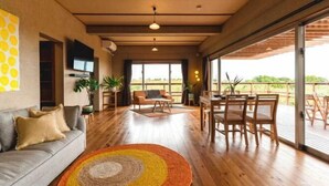 Een ruime woonkamer met houten vloer, een bank, een eettafel met stoelen en grote ramen met uitzicht op de omgeving.