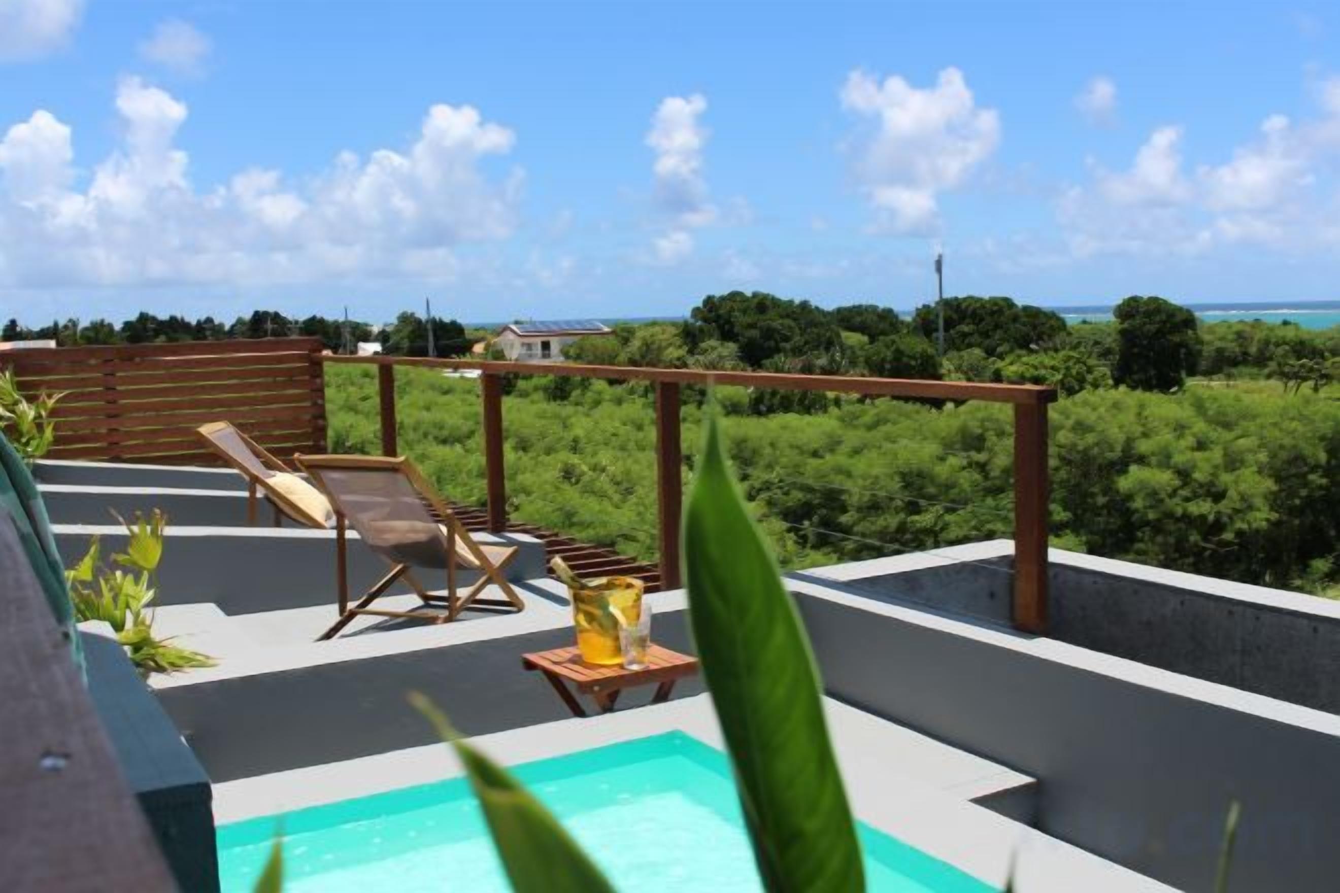 A rooftop pool area with lounge chairs and a small table, surrounded by greenery and a view of the ocean.