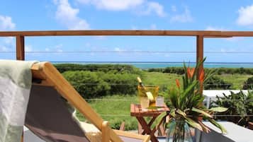 A balcony with a view of the ocean, a wooden chair, a small table with a vase of flowers, and a wooden railing.