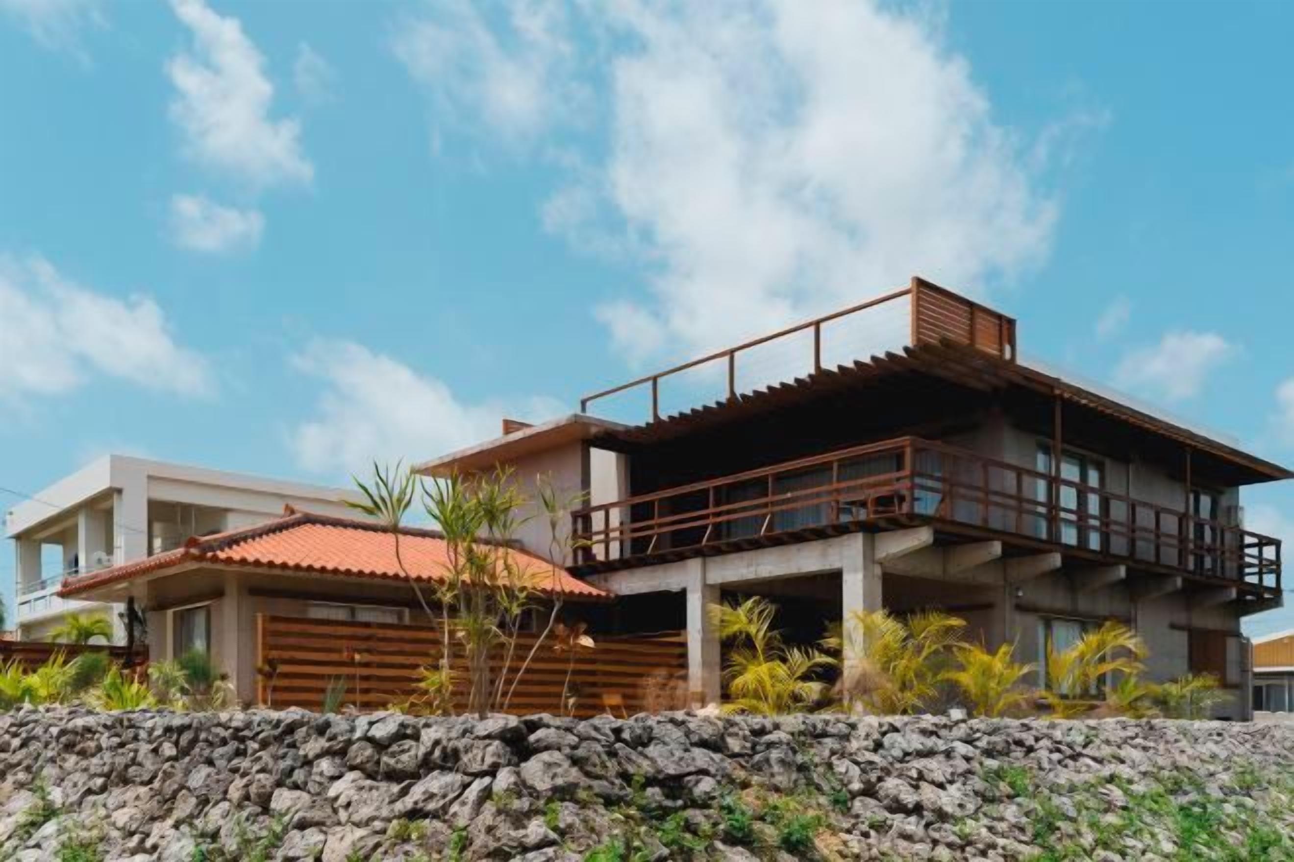 A modern two-story house with a balcony, a stone wall in the foreground, and a clear sky.