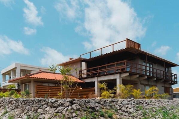 A modern two-story house with a balcony, a stone wall in the foreground, and a clear sky.