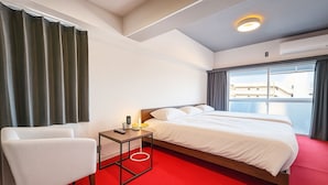 A hotel room with a bed, a chair, a small table with a lamp, and a view of buildings through the window.