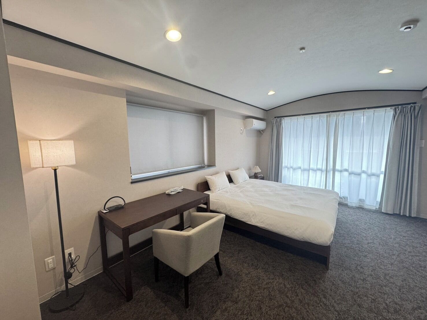 A modern hotel room with a large bed, a desk, a chair, a lamp, and a window with curtains.