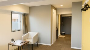 A hotel room with a single white chair, a small table with a remote control, a window with curtains, and a door leading to a hallway.