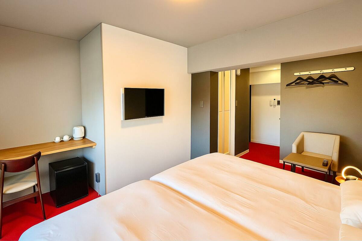 A hotel room with a bed, a desk, a chair, a TV, a mini-fridge, and a coat rack.