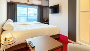 A hotel room with a bed, a TV mounted on the wall, a desk with a lamp, and a red carpet.