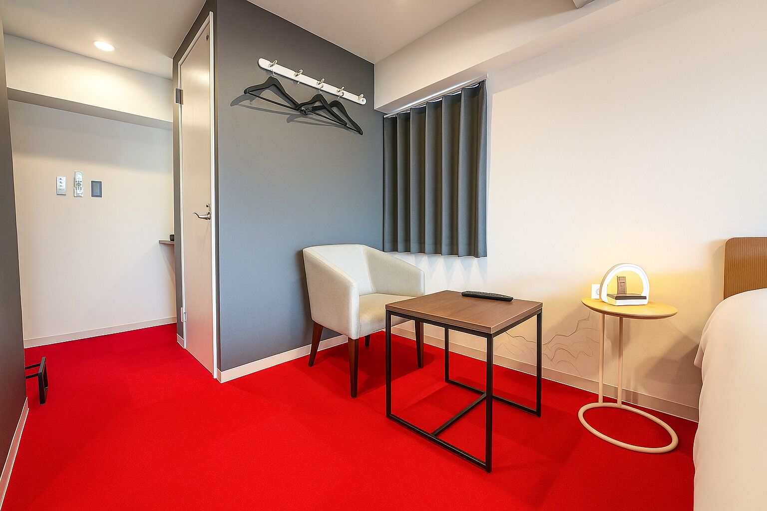 Ein Hotelzimmer mit rotem Teppich, einem weißen Sessel, einem kleinen Tisch, einem Bett, einem Nachttisch mit Lampe und einem Kleiderständer mit Kleiderbügeln.