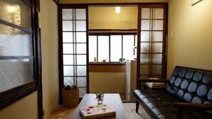 Una habitación japonesa tradicional con suelo de tatami, una mesa baja con un libro y un florero, un sofá de cuero negro y puertas correderas que dan a una zona de cocina.