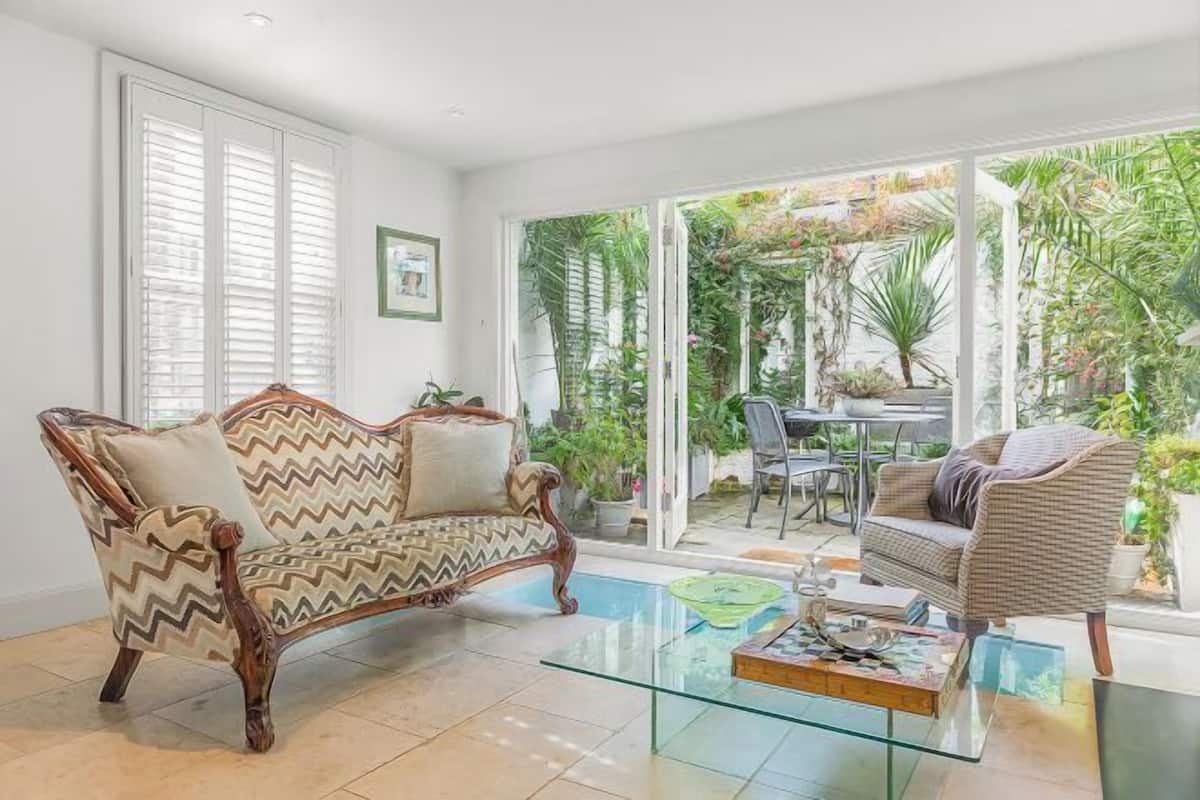 A living room with a sofa, a glass coffee table, and a view of a garden through large windows.