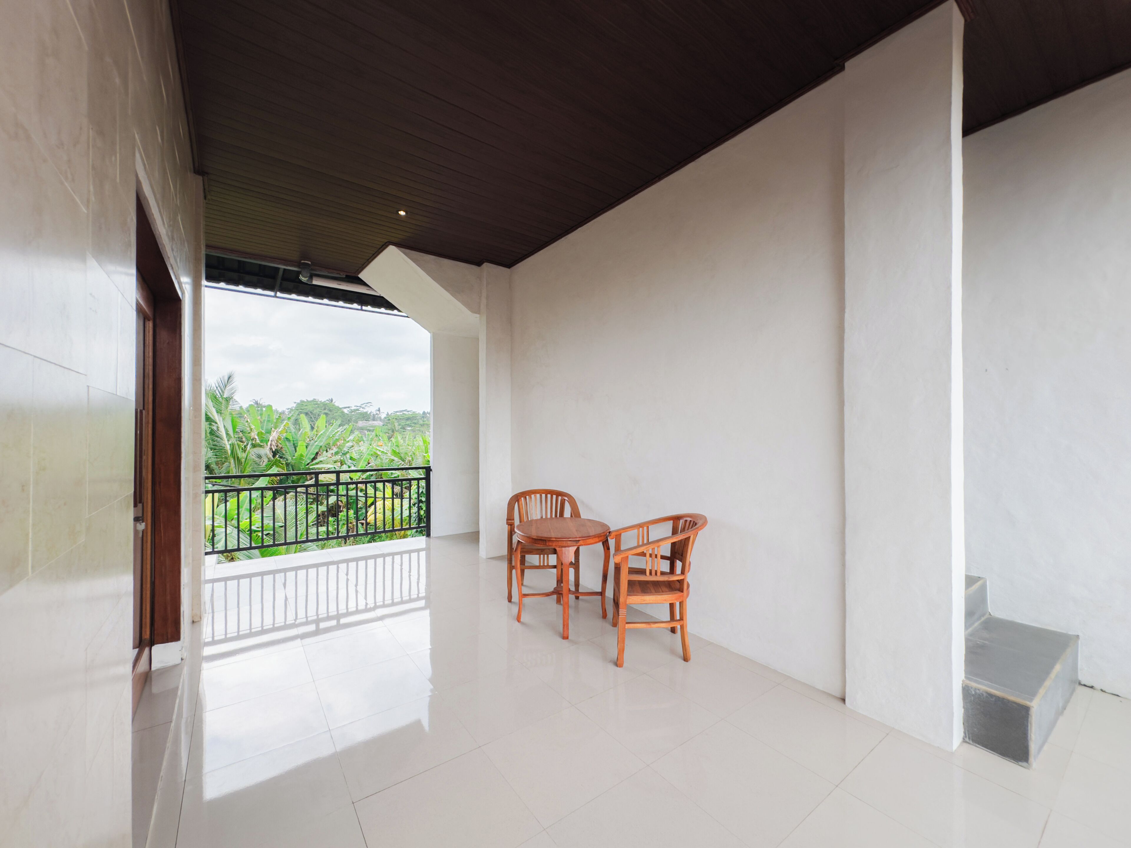 A spacious balcony with a wooden table and chairs, a view of greenery, and a white tiled floor.