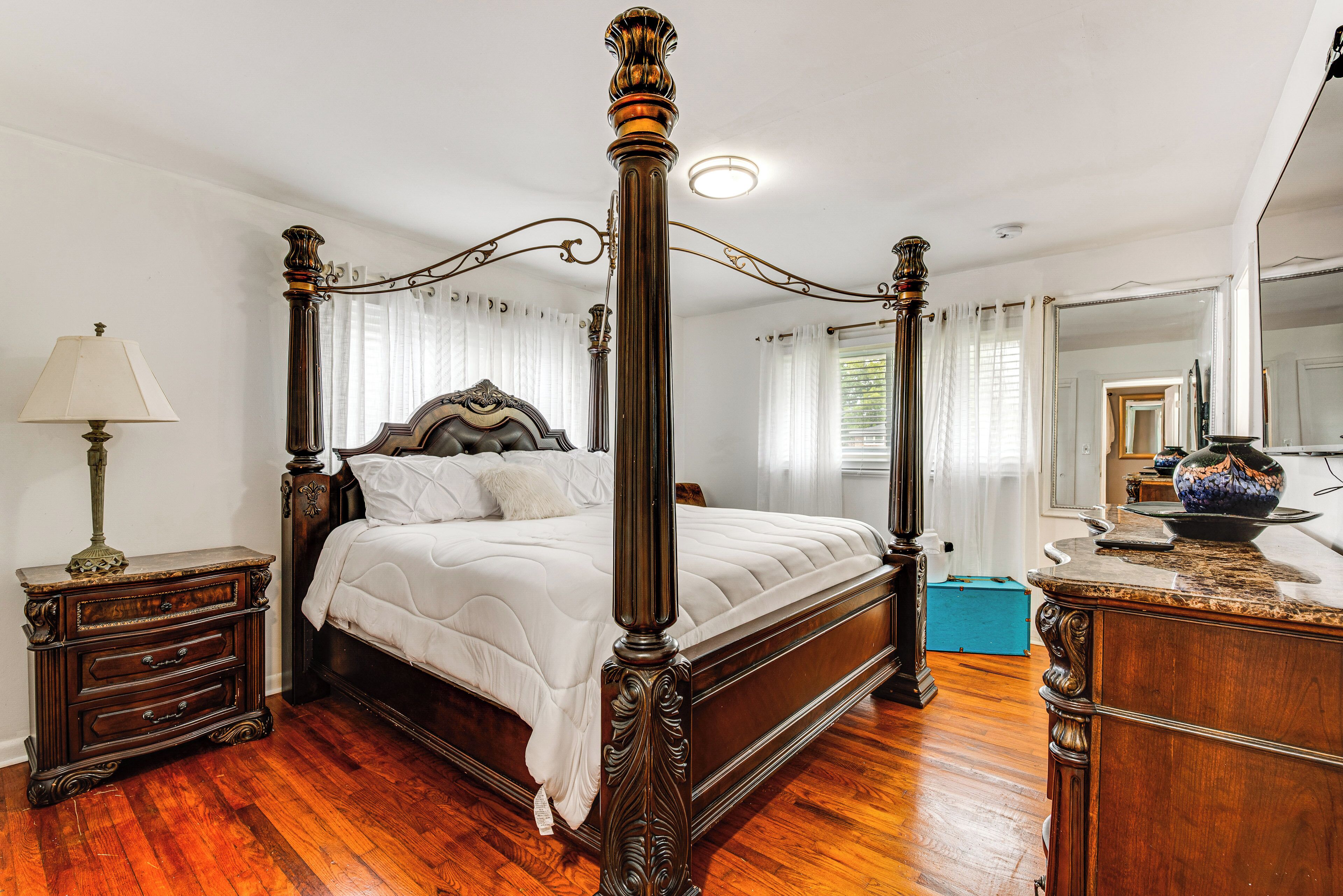 A four-poster bed with a wooden frame and white bedding, a nightstand with a lamp, and a dresser with a vase.