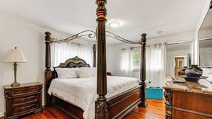 A four-poster bed with a wooden frame and white bedding, a nightstand with a lamp, and a dresser with a vase.
