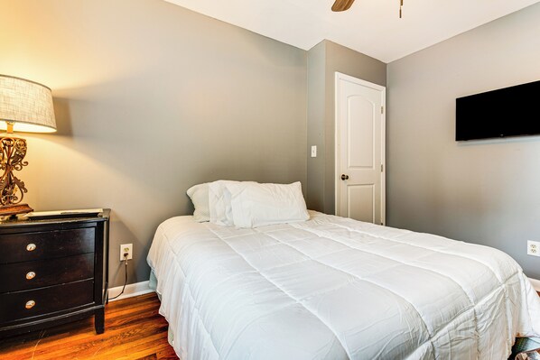 A bedroom with a bed, a dresser, a lamp, a door, and a TV mounted on the wall.
