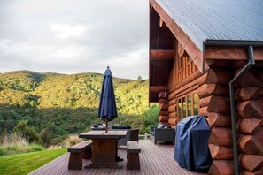 A wooden deck with a table and chairs, a covered grill, and a view of a forested hillside. - Cascade Creek Retreat (Circle Hill)