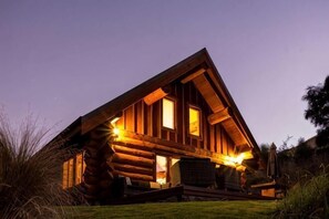 Une cabane en rondins dotée d’une véranda, de grandes fenêtres et d’un intérieur bien éclairé.
