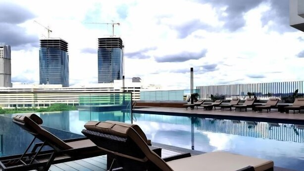 A rooftop pool with lounge chairs, a glass barrier, and a view of modern buildings.