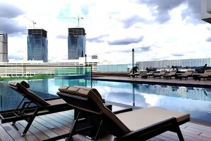 A rooftop pool with lounge chairs, a glass barrier, and a view of modern buildings. - Cityview MidValley Southkey 2BR+2FREE 16FL BY NATOL (Plentong)