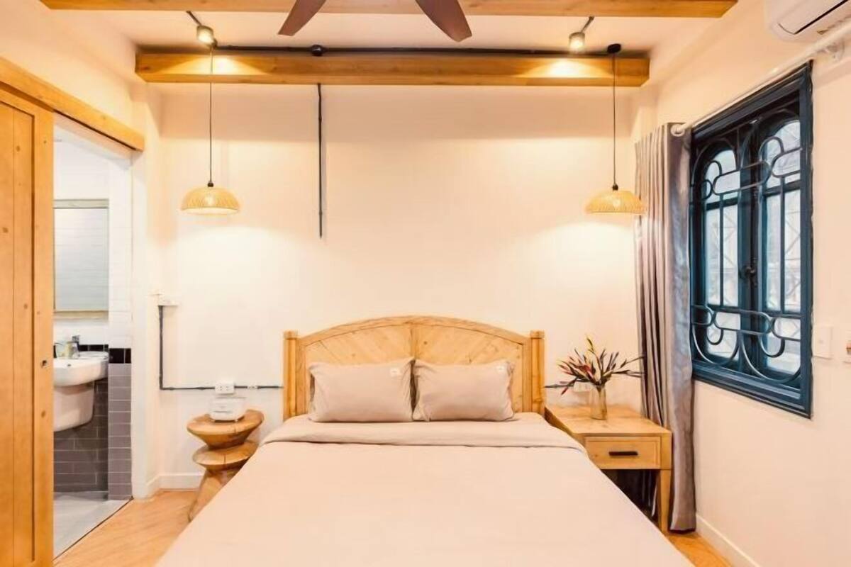 Un dormitorio con cama de madera, mesitas de noche, una ventana con cortinas y un baño visible a través de una puerta abierta.