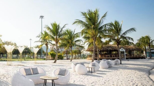 A beachside lounge with white cushioned seating and small tables, surrounded by palm trees and a building with a thatched roof. - Yichen Apartment - Vinhomes Ocean Park Ha Noi (Hanoi)