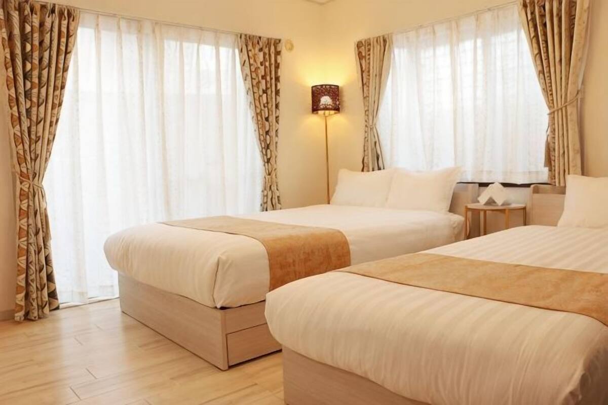 A hotel room with two beds, a bedside table, a lamp, and curtains.