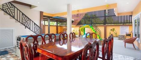 Un comedor amplio con una gran mesa de madera y sillas tapizadas en rojo, una escalera y un área de juegos colorida al fondo.