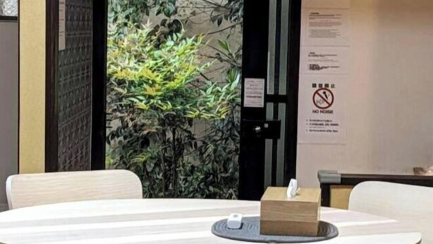 Une table ronde avec deux chaises, une boîte de mouchoirs et un petit appareil électronique posé sur la table. Il y a une porte avec une pancarte accrochée au mur à côté.