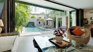 A modern living room with a glass coffee table, a wicker chair, and a view of an outdoor swimming pool. - Villa Chili in Dalung (Bali)