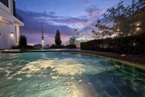 Uma piscina com água cristalina, rodeada por um jardim ajardinado com árvores e arbustos, e um edifício moderno ao fundo.