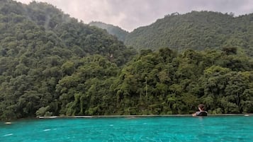 A person sitting in a clear blue pool with a forested hill in the background.