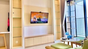 A modern living room with a flat-screen TV mounted on the wall, a round table with a remote control, and a green sofa. - LukaHomes - Dcapitale/1BR+/City view (Hanoi)