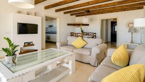 Ein Wohnzimmer mit einem Glastisch, zwei Sofas mit gelben Kissen, einem an der Wand montierten Fernseher und freiliegenden Holzbalken an der Decke.