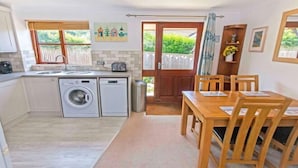 A compact kitchen with a washing machine, a sink, and a dining area with wooden chairs and a table. - Cockleshell Croyde Sleeps 4 By the Seaside (Croyde)