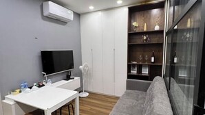 Un salon moderne avec un bureau blanc, un canapé gris et un téléviseur. On y trouve également une armoire blanche, un ventilateur et une étagère avec des bouteilles et des verres.