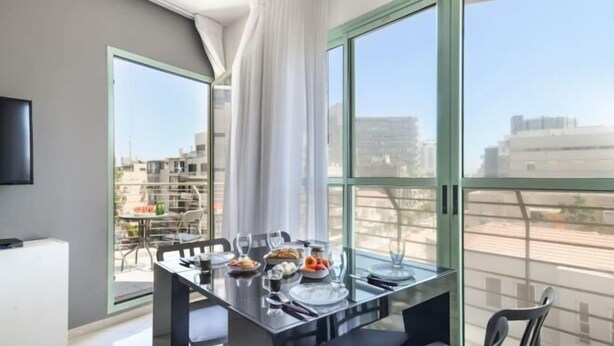 A modern dining area with a glass table, chairs, and a view of buildings through large windows. - Elegant Sea-View Apt w Mamad by Sea N' Rent (Tel Aviv)