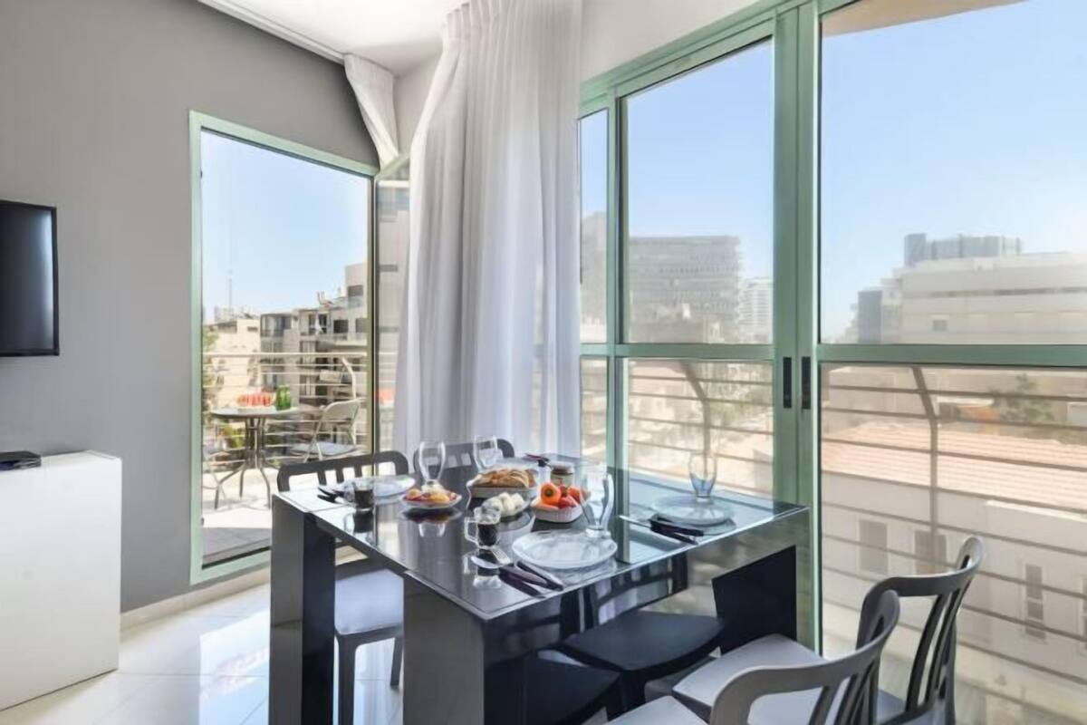 Un comedor moderno con una mesa de vidrio, sillas y vistas a edificios a través de amplios ventanales.