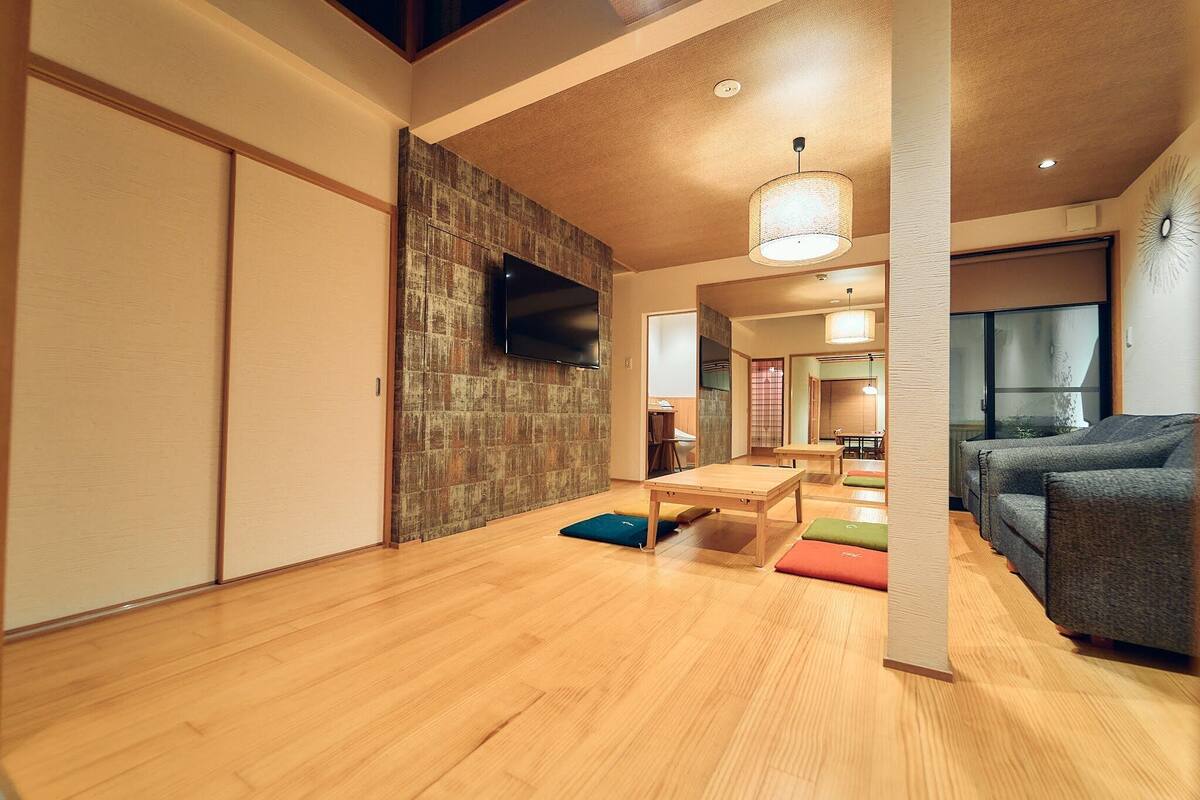 A modern living room with a wooden floor, a flat-screen TV mounted on a textured wall, a dining table with cushions, and a grey sofa.