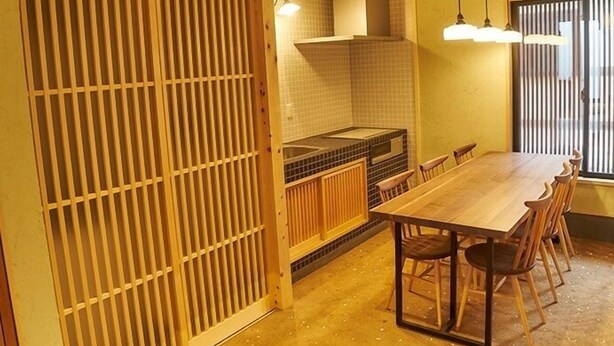 Une pièce de style japonais traditionnel, avec un sol recouvert de tatamis, une basse table en bois entourée de chaises, un coin cuisine équipé d’une cuisinière et d’un four à micro-ondes, et une porte coulissante à lattes verticales.